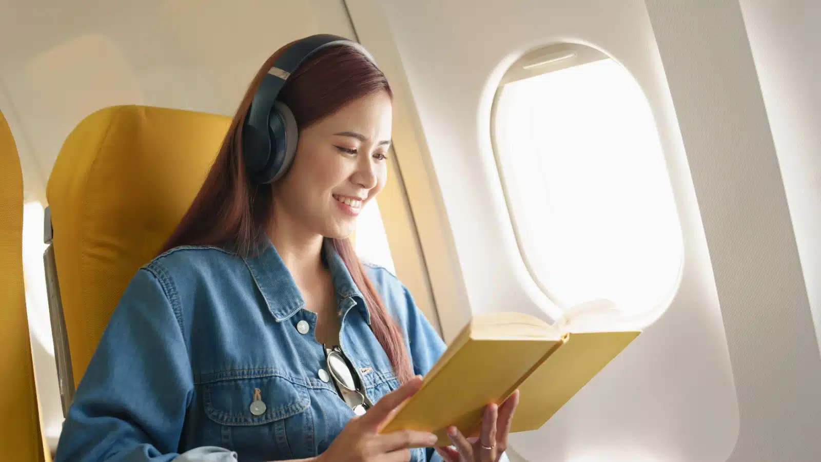 A woman wears headphones and reads a book while flying.