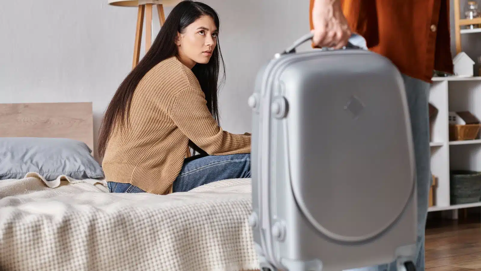 A sad woman sitting on the bed as her partner leaves with a suitcase. He booked a trip without telling his girlfriend.