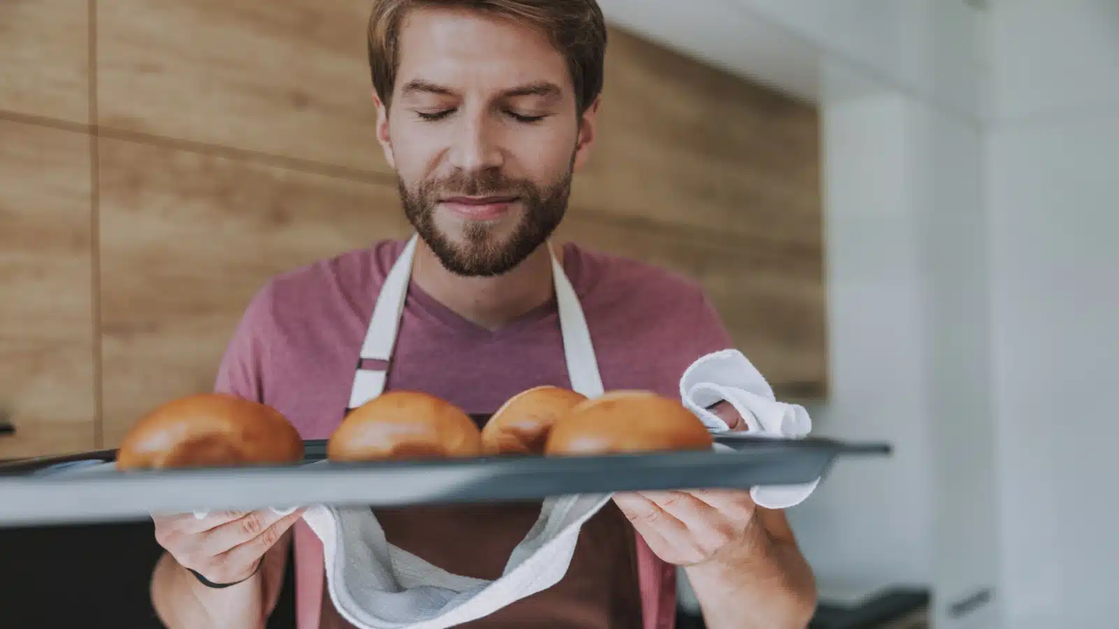 A man smiles as he smells the tray of freshly baked bread he is holding.