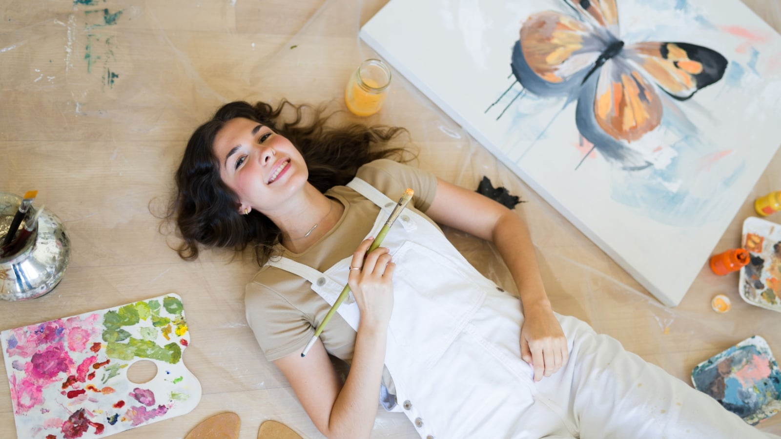 A smiling artist lies on the floor in her studio surrounded by art supplies to represent gifts for artists.