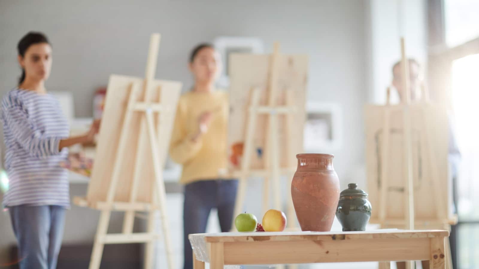 Art class students stand at easels, painting a still life scene. 