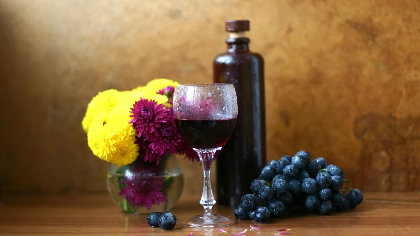 Items set up for a still life painting include flowers in a vase, grapes, a glass of wine, and a bottle. The items showcase contrasting textures, from the smooth bottle, to the puffy flowers. 