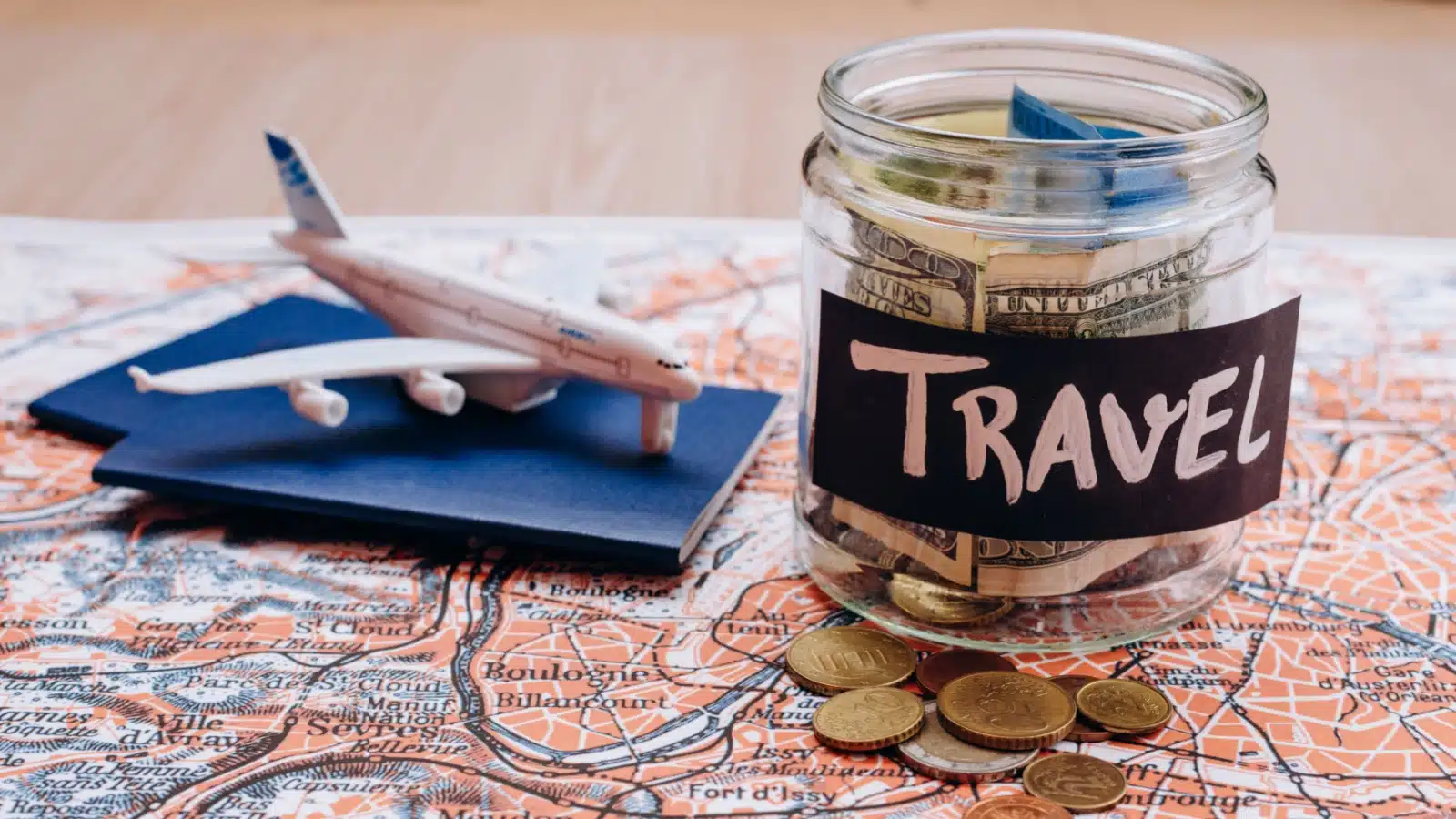 A money jar labelled "travel" sits on a map next to a model airplane and passport to represent a travel budget.