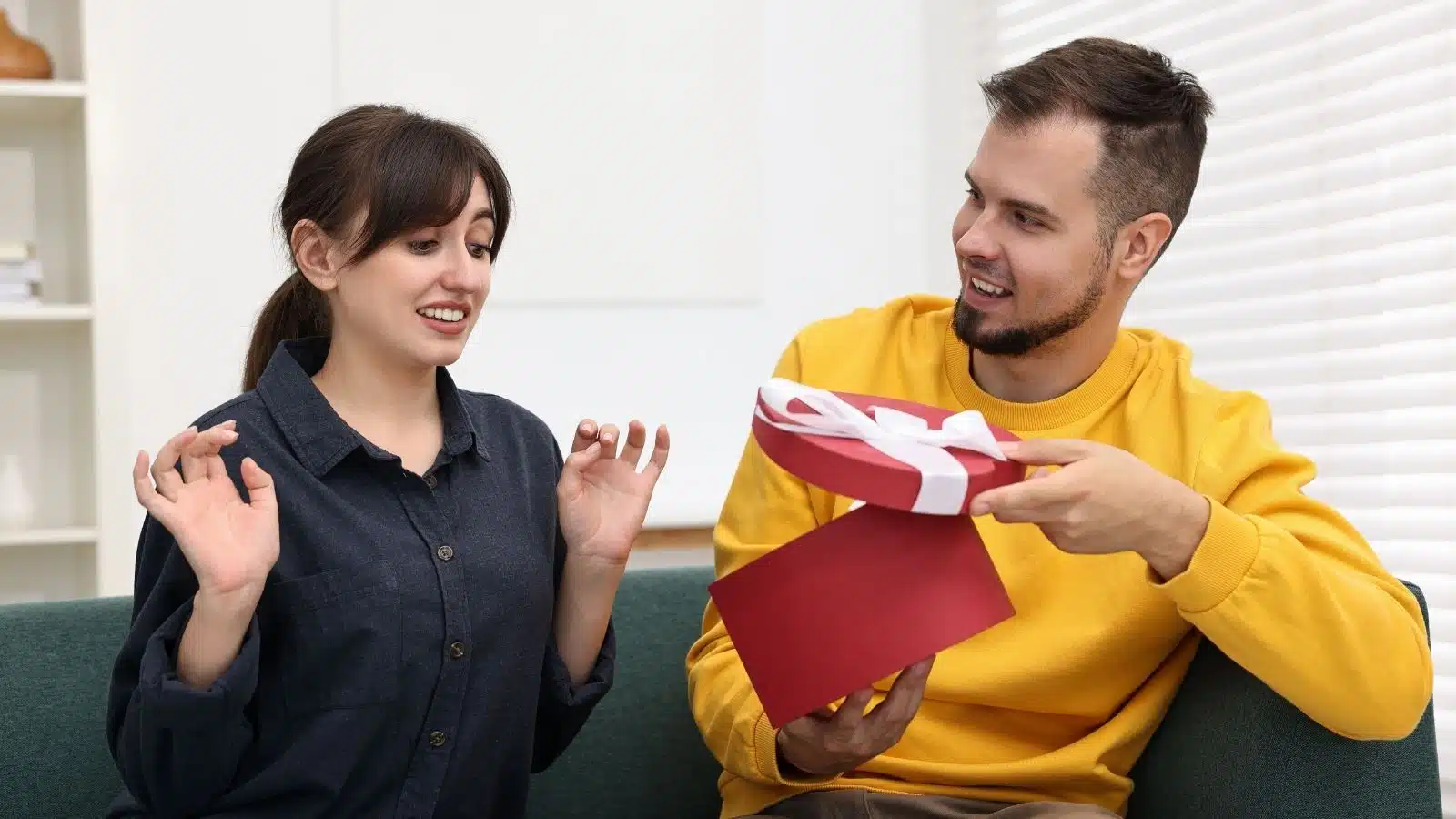 A happy man presents a gift to his girlfriend, who is trying to look surprised but clearly hates it. The photo showcases a bad gift giver boyfriend.
