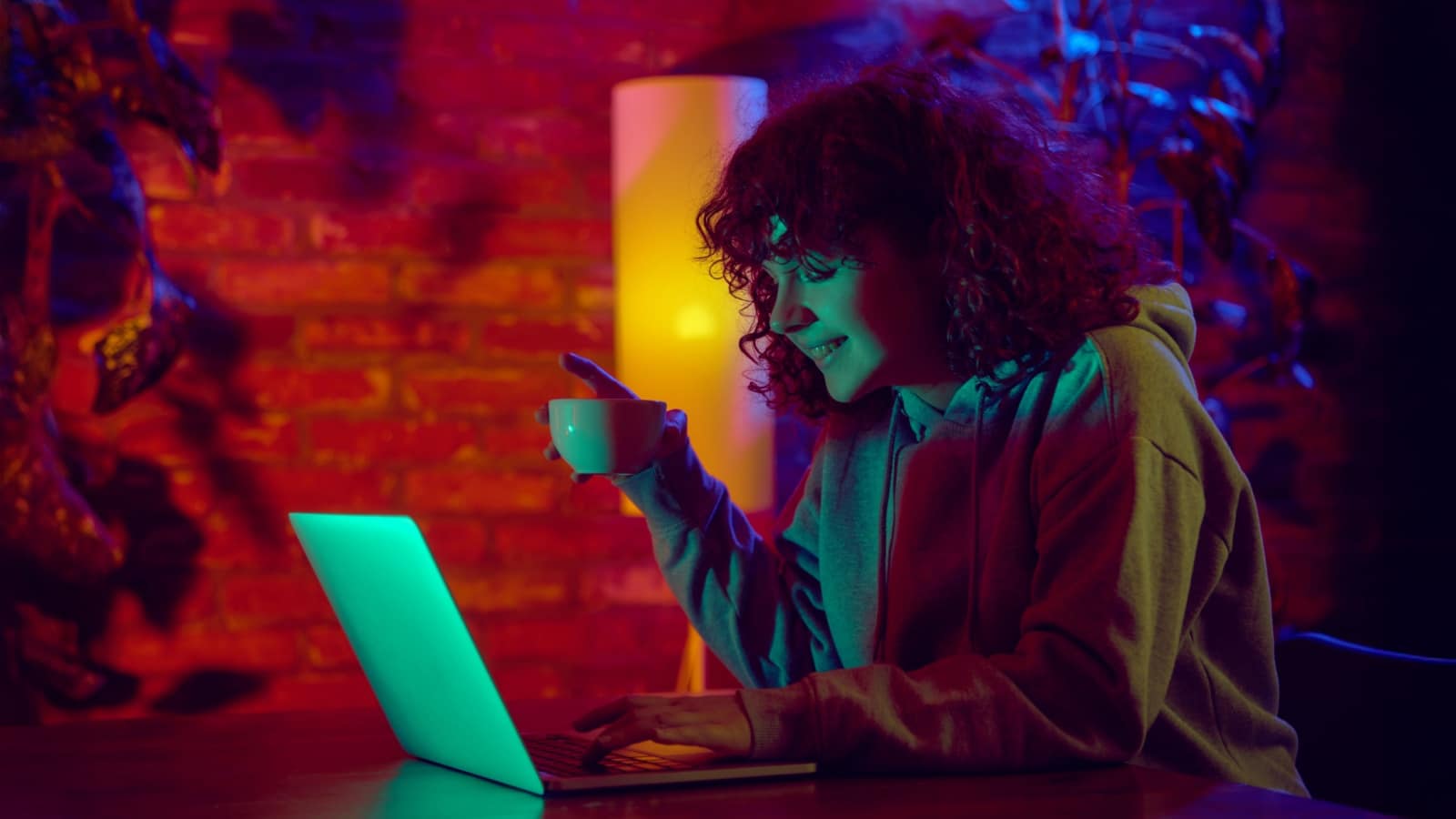 A happy woman uses her laptop in a dark yet colorful space to represent creative writing.