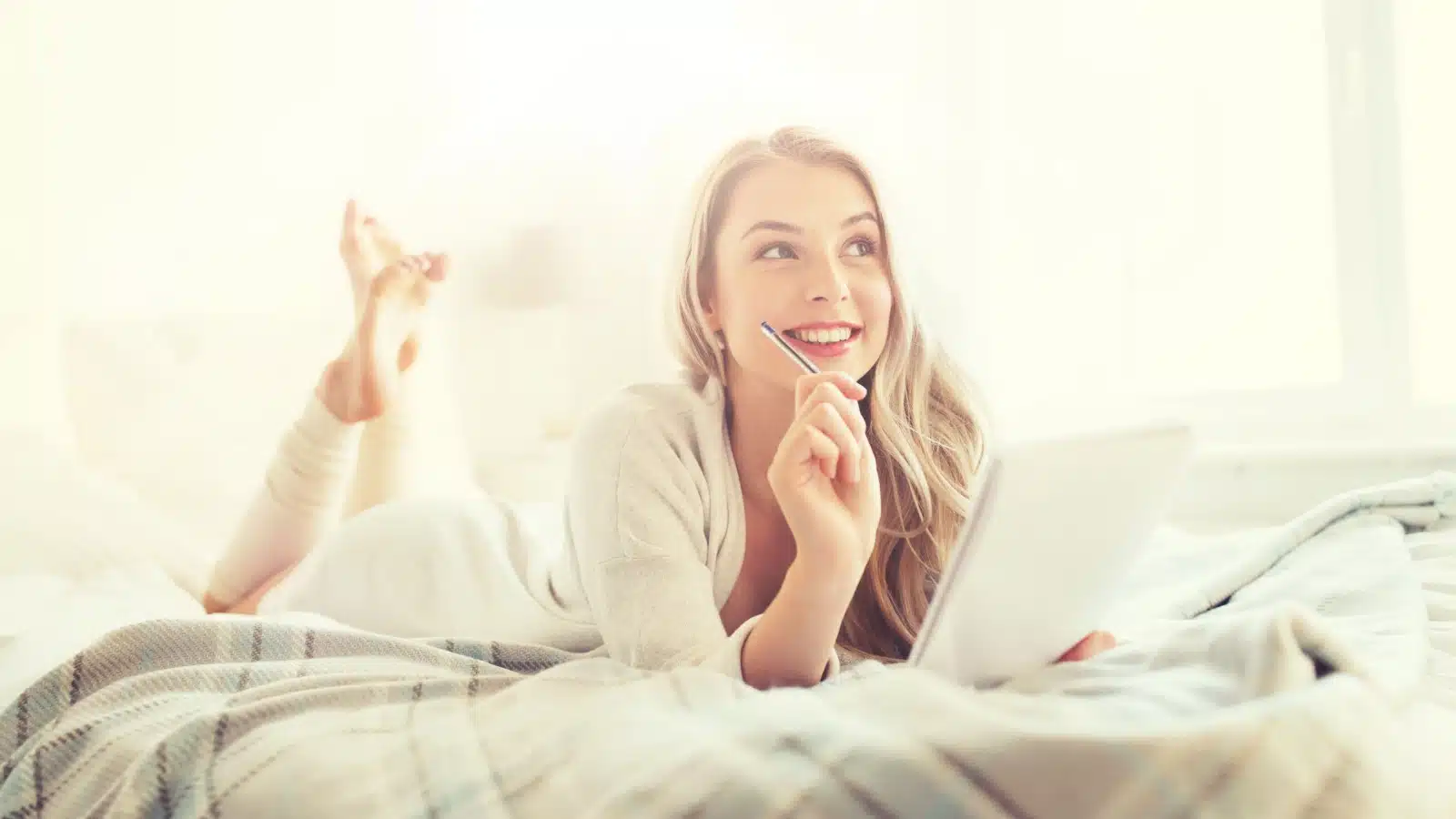 A smiling woman lies on her bed with her notepad and a pen, to represent fun lists to make.