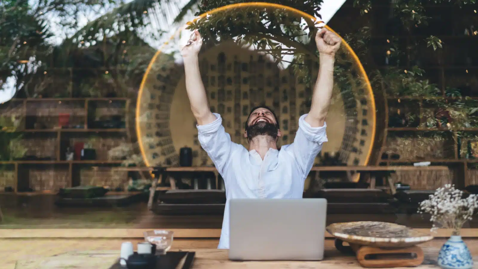 A man celebrates his accomplishments. He's sitting in front of his laptop cheering.