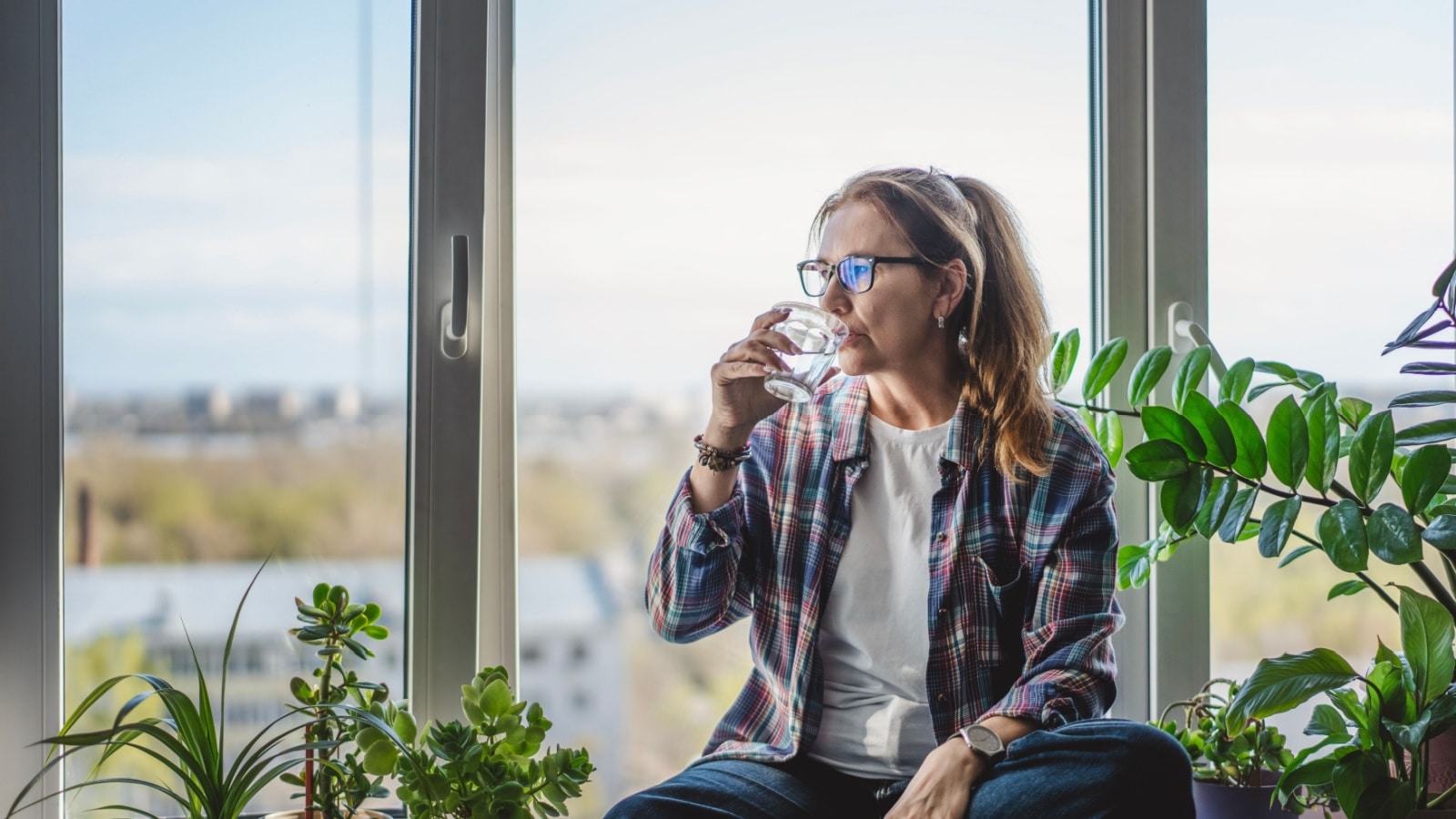 A woman sips water while sitting in front of giant glass windows of the house she's watching. 