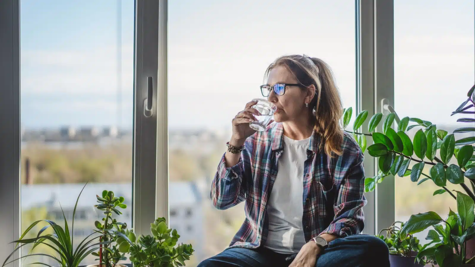 A woman sips water while sitting in front of giant glass windows of the house she's watching.