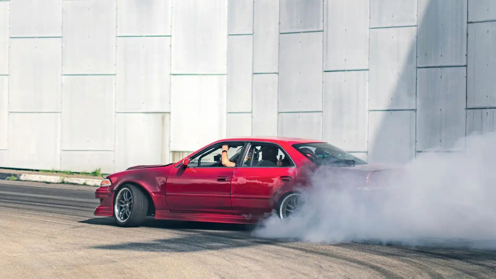 A red car drifts on the pavement with smoke coming out of the exterior. It's probably loud.