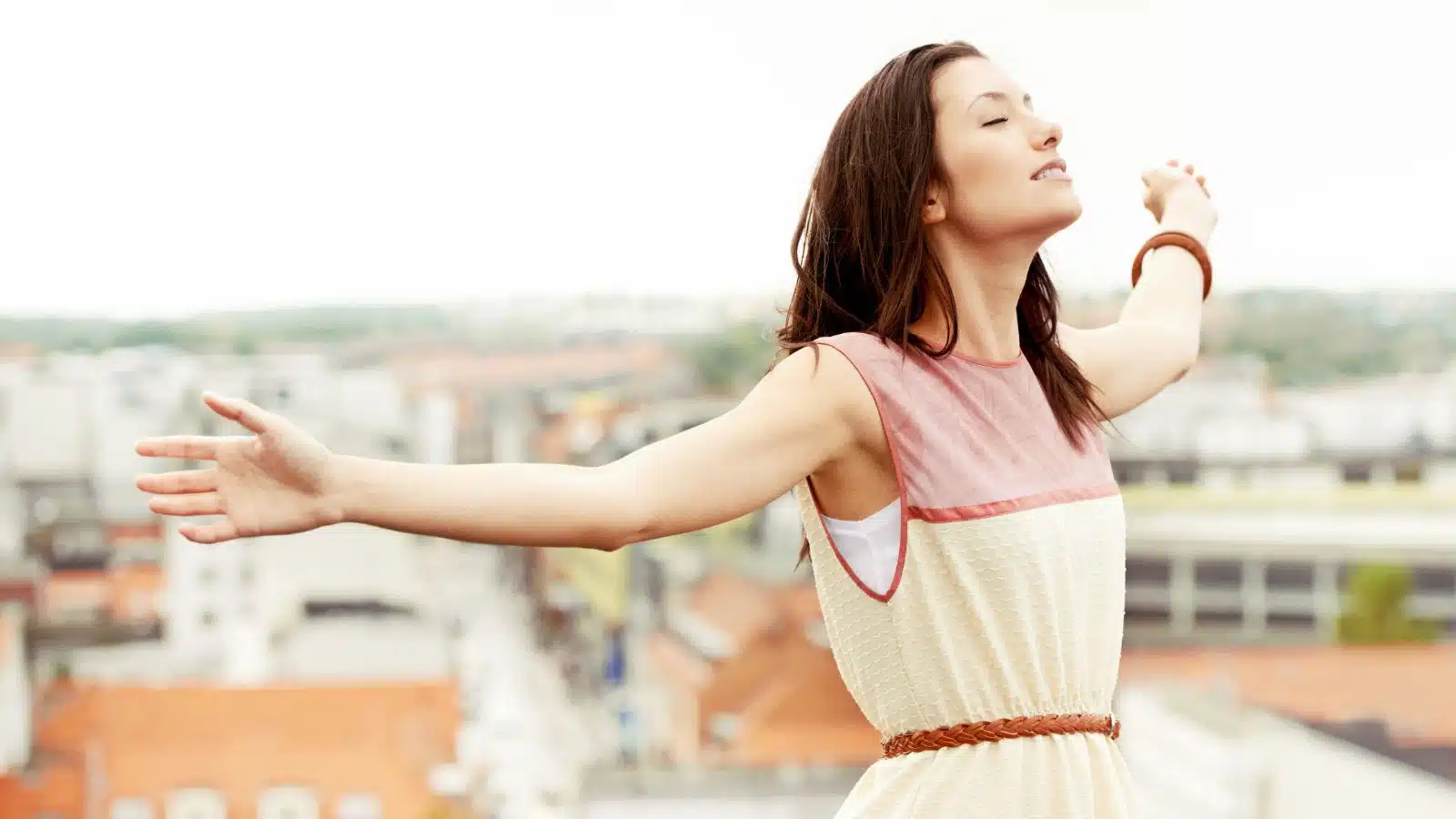 A woman stands on a roof top with her arms spread wide, breathing in deep and relaxing as she lets go of trivial things and lets the stress melt away.