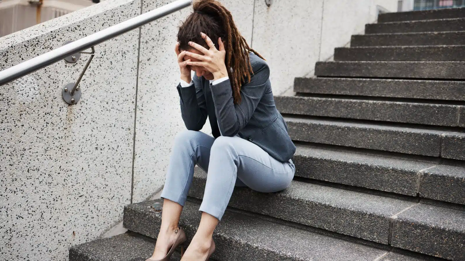 An upset scared woman sits alone on concrete stairs with her head in her hands.