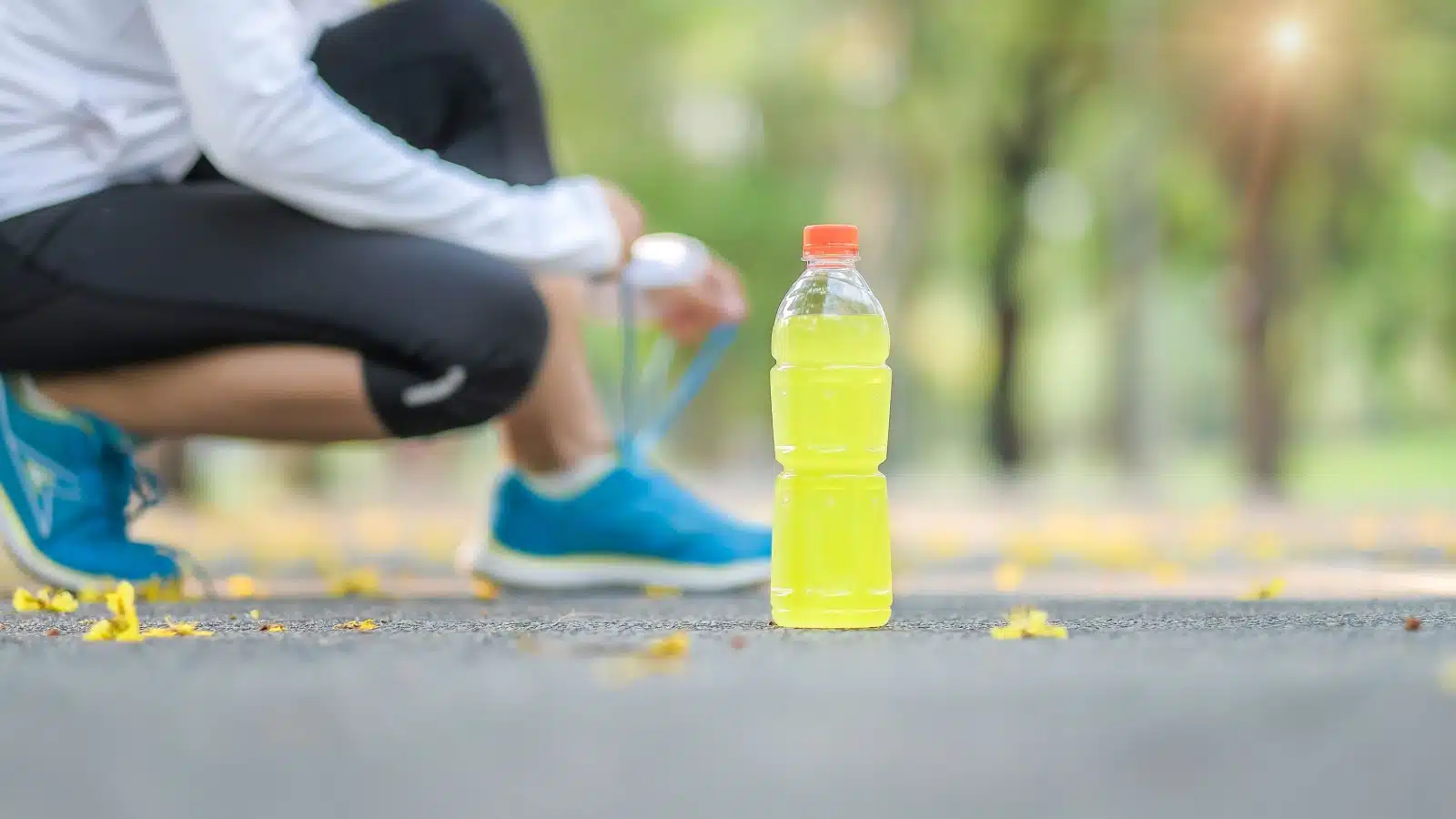 A yellow sports drink rests on the sidewalk while a runner ties her shoes in the background.