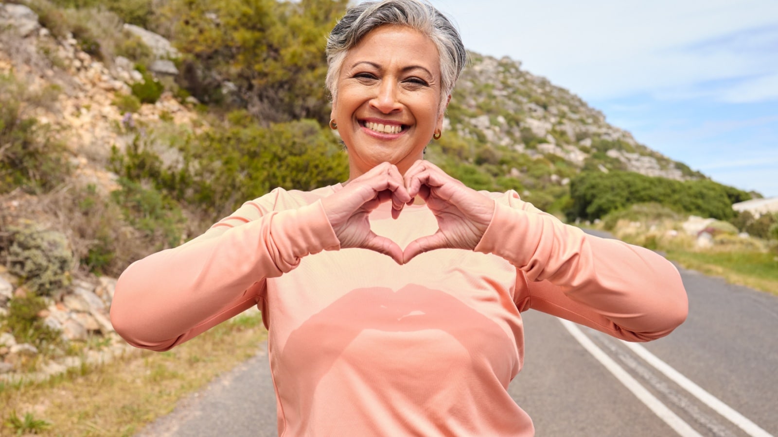 A smiling middle aged women in the middle of a scenic mountain road making a heart sign with her hands to represent focus on the good.