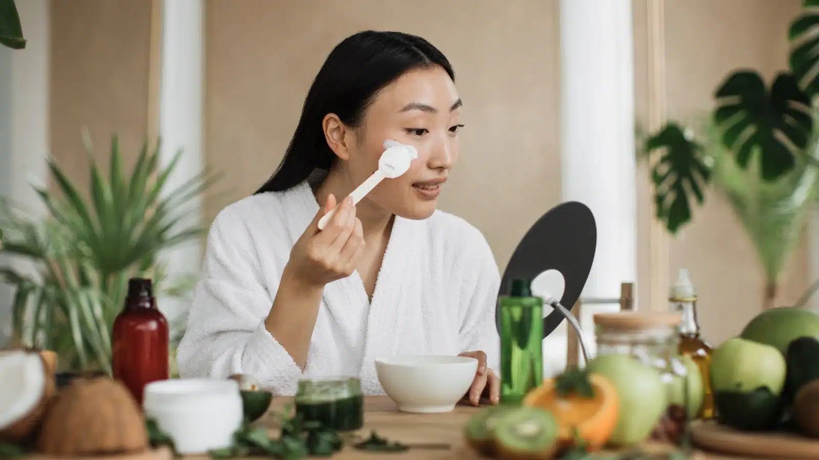 A woman applies a homemade lotion to her face. The lotion ingredients are on the table in front of her.