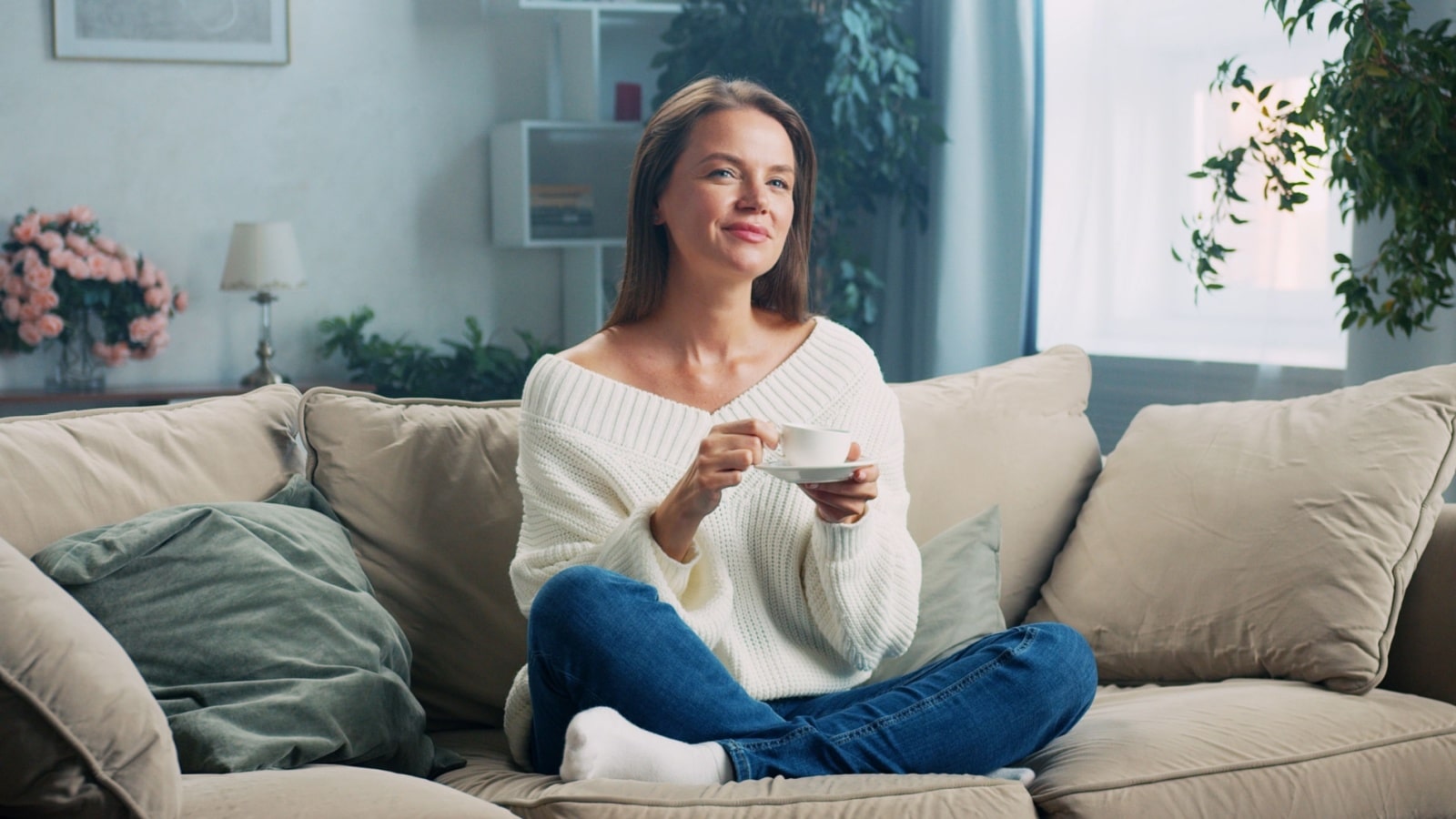 A woman sits happily on her couch, enjoying a warm beverage, to represent how to savor the moment.