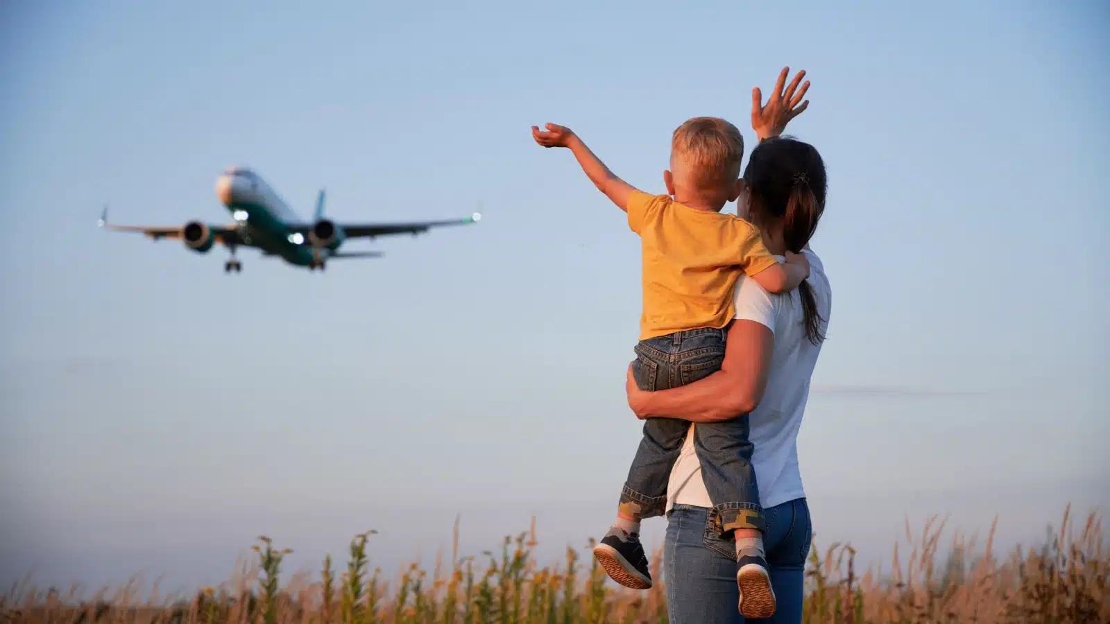 A woman holds her young son while they both wave goodbye to an airplane flying overhead.