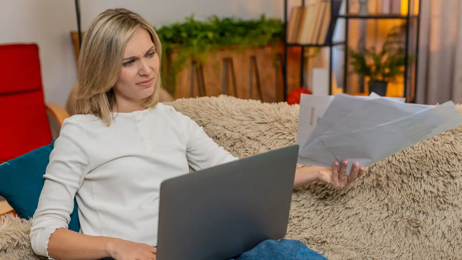 An annoyed middle aged woman holds papers while working on her laptop.