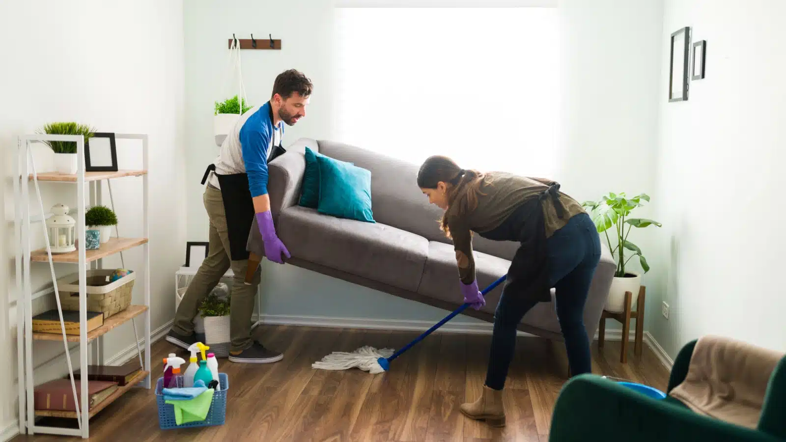 A woman cleans under a couch while her husband lifts it.