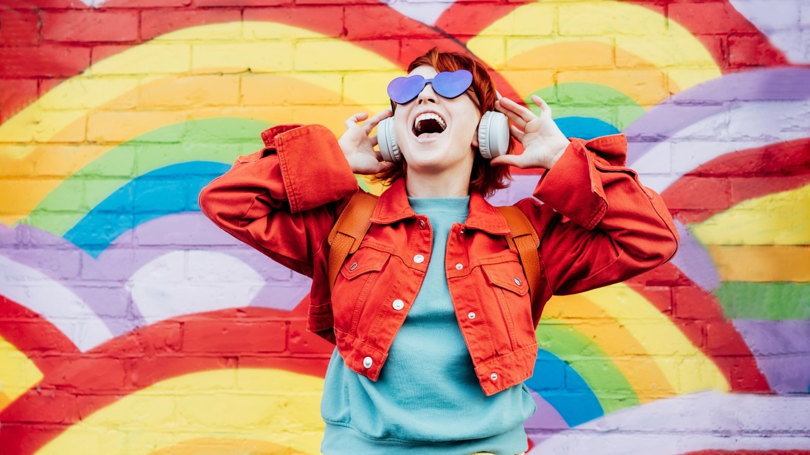 A happy woman stands in front of a colorful mural on a brick wall. 