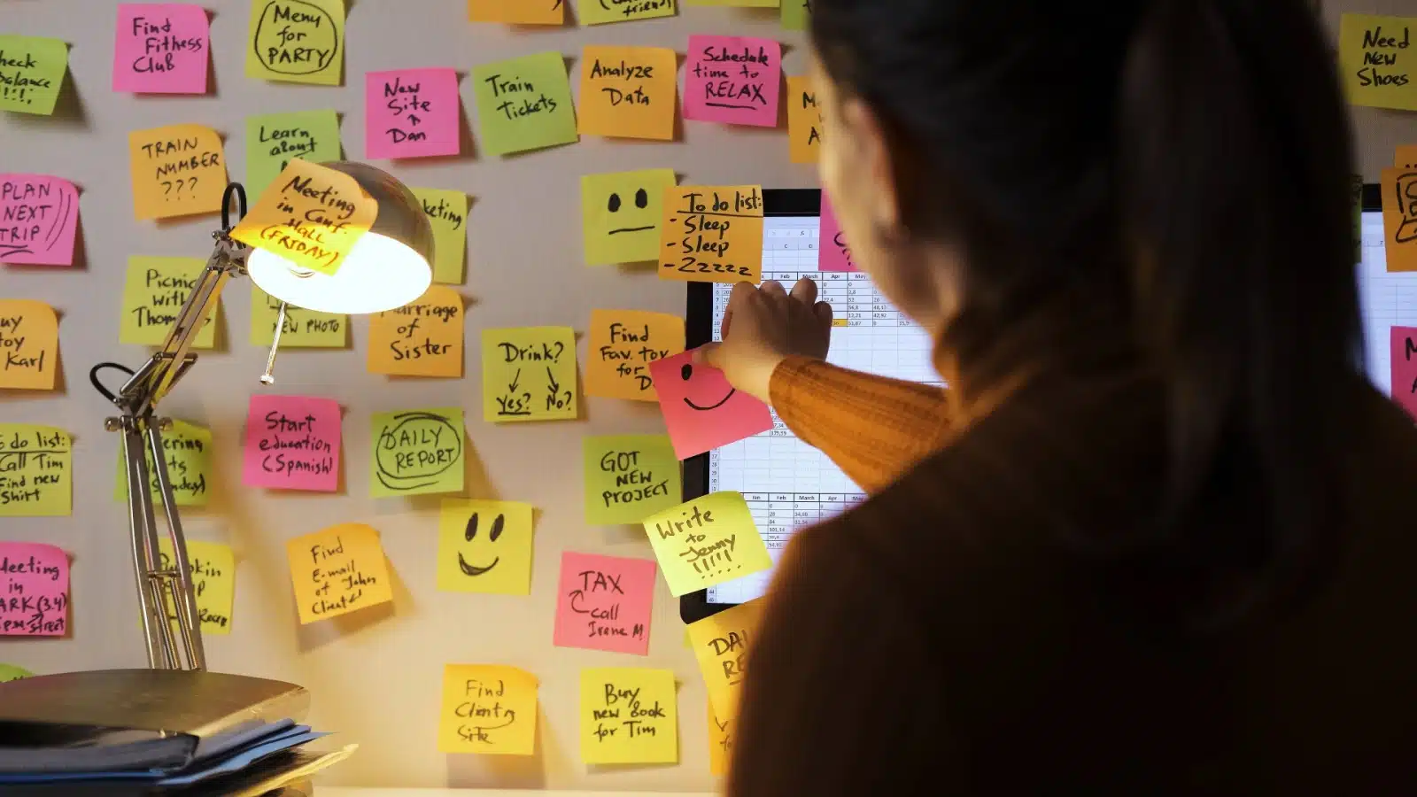 A woman places another post it note on her wall, reminding herself to do something. Her busy wall of post-its represents the mental load.