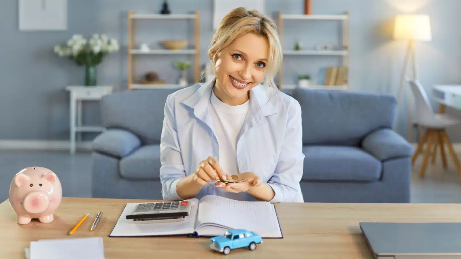 A model car rests on a table in front of a woman who's smiling while putting money in a piggy bank, to represent saving money on car payments and car insurance.
