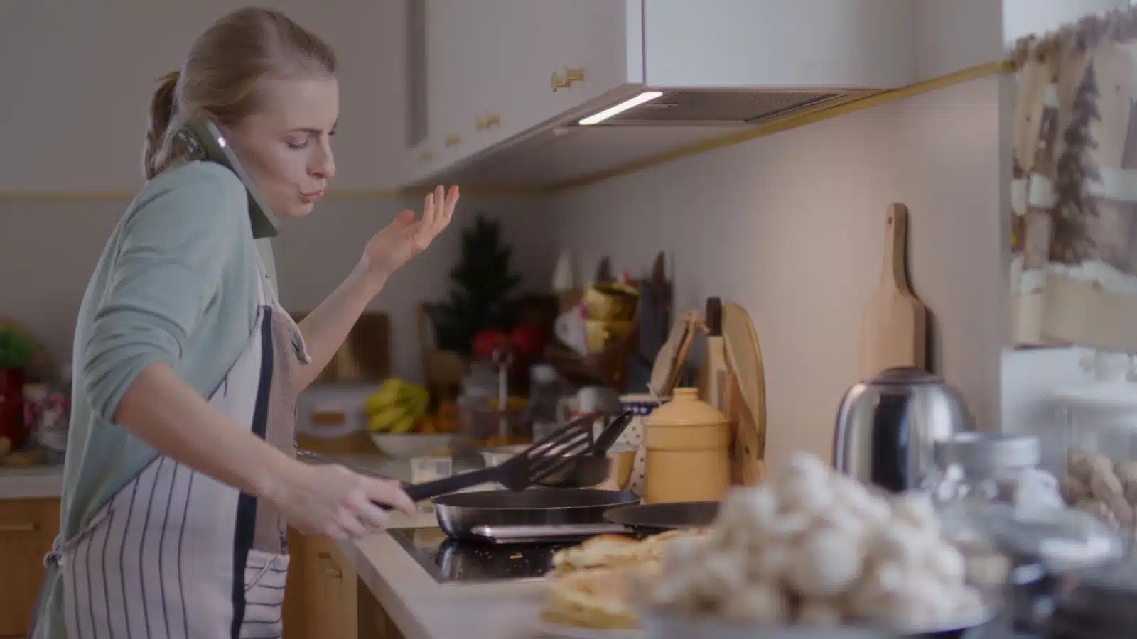 A stressed woman talks on the phone while cooking.
