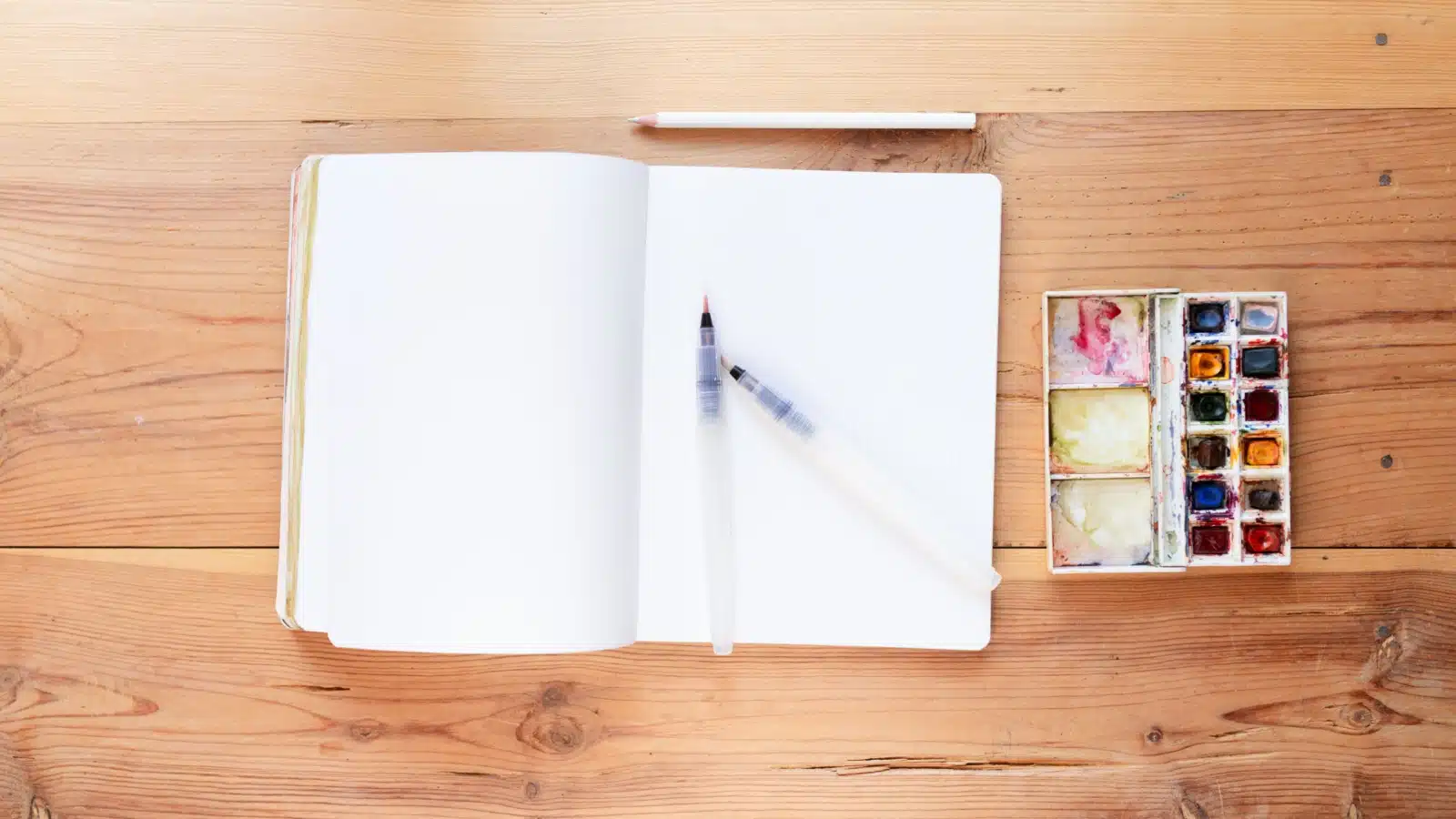 A blank art journal next to a set of watercolor paints.