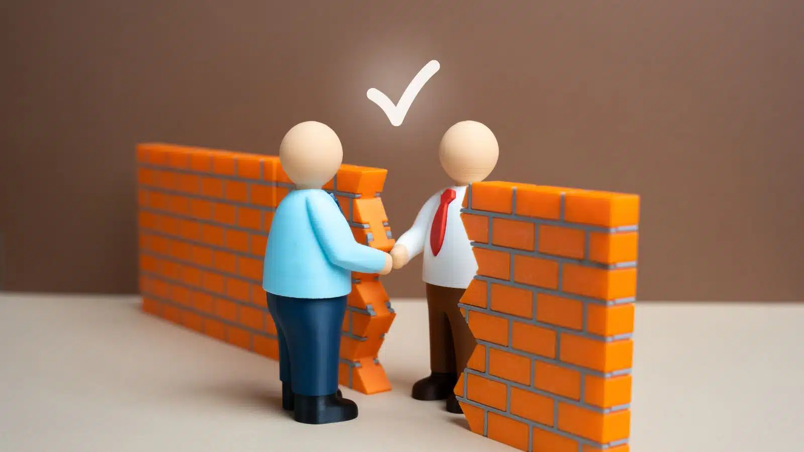 Two model businessmen shake hands through a break in a brick wall, representing compromise.