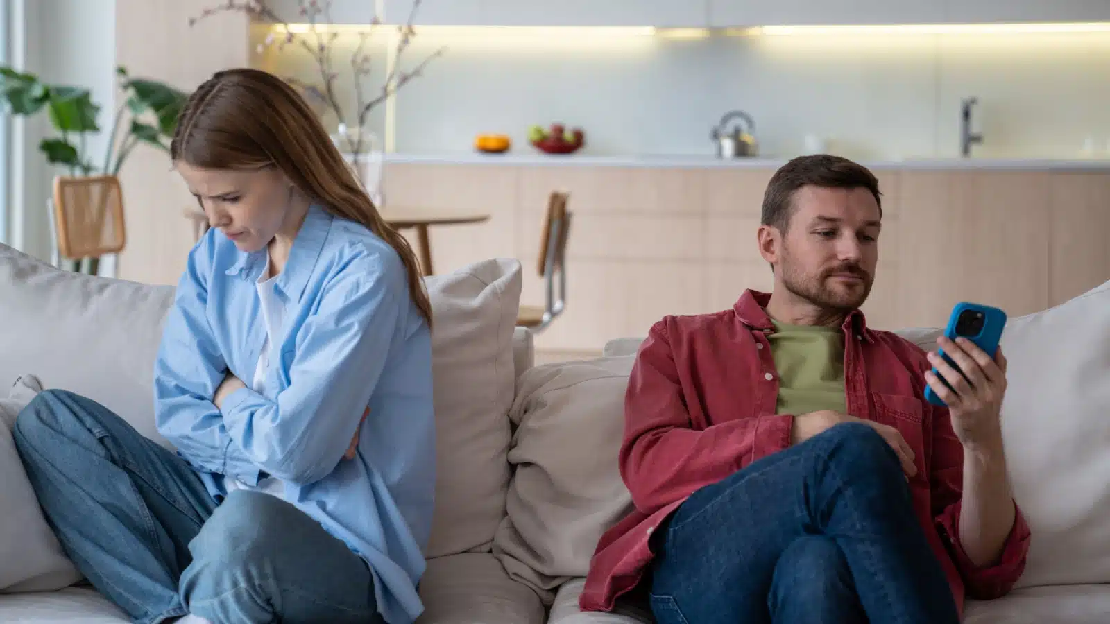 A sad wife sits on the couch next to her husband, who happily plays on his phone because he doesn't care.