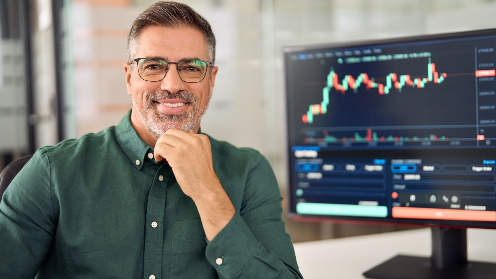 A smiling middle aged man in front of his computer showing an investment graph to represent secret millionaire habits you can copy.