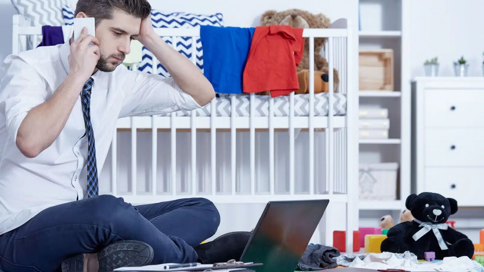 An overwhelmed father tries to work while sitting next to a baby's crib.