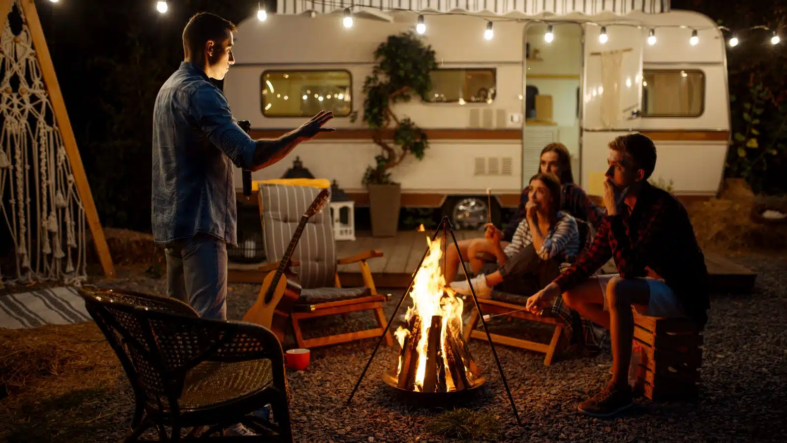A man tells campfire stories to his friends. They're at a campsite, with a small trailer, sitting around the fire.