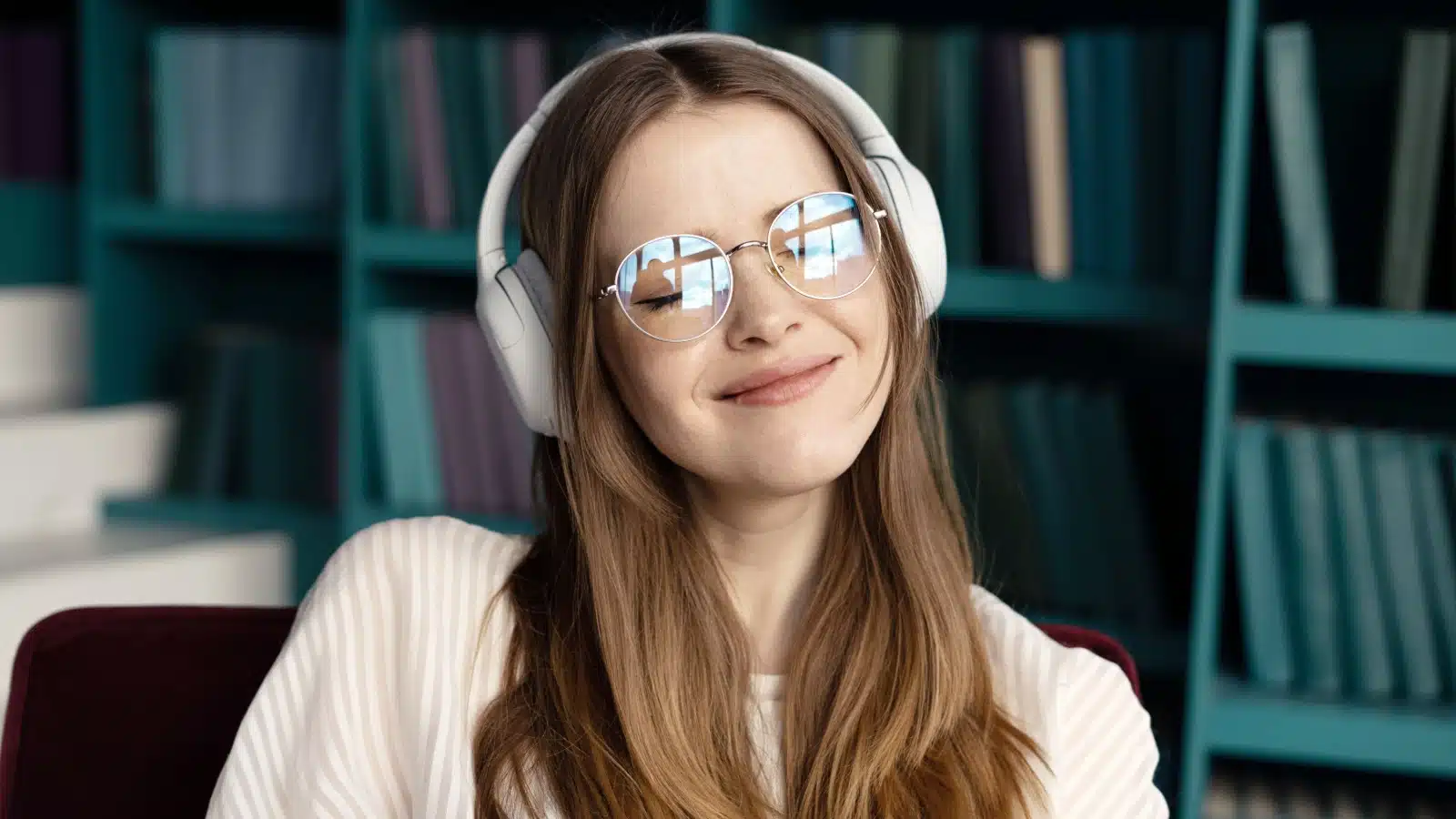 A girl in a library listens to music on her headphones. She looks hopeful.