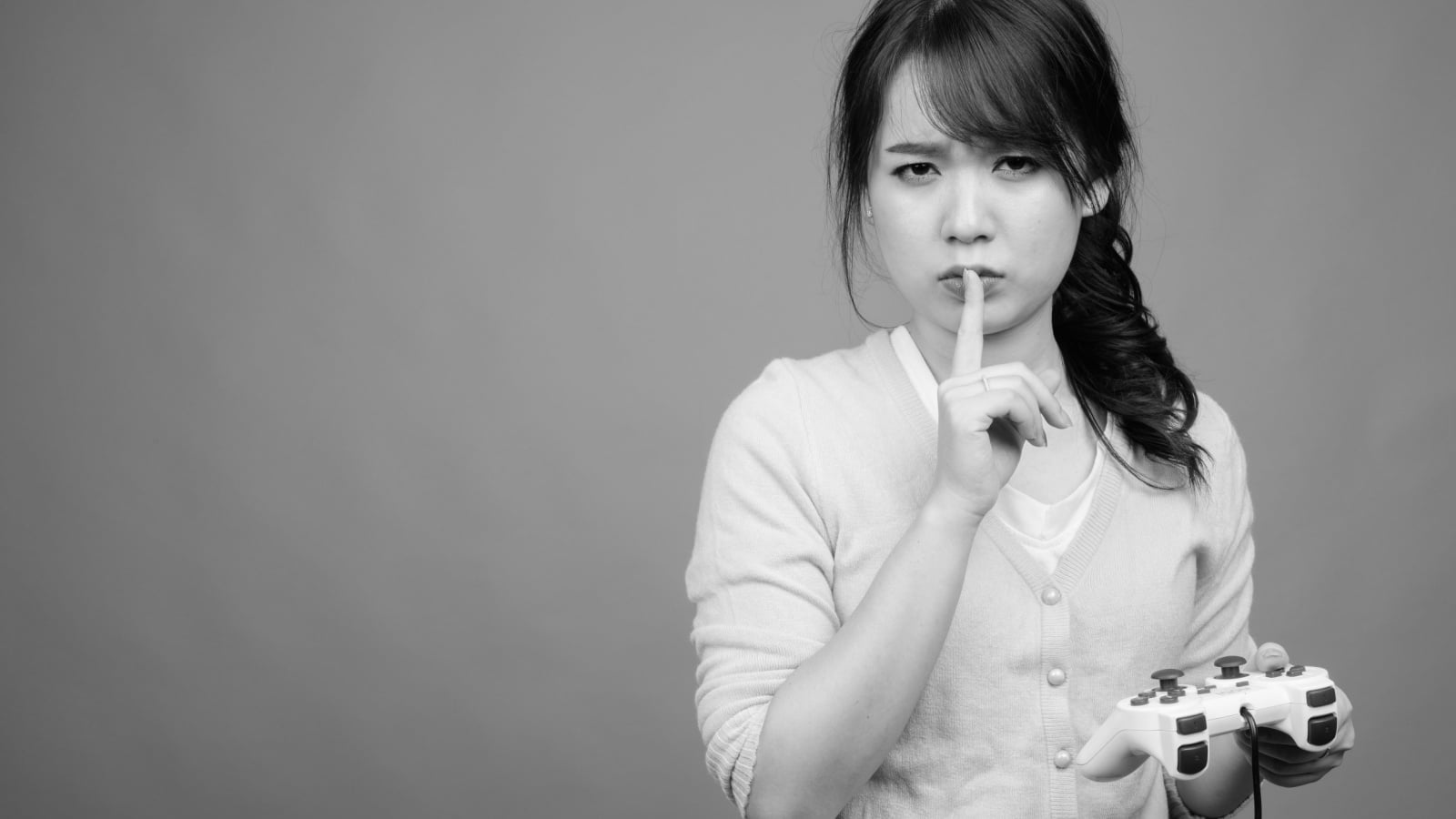 Black and white photo of a woman holding a video game controller while making the silence motion with a finger over her mouth to represent sexism in gaming.