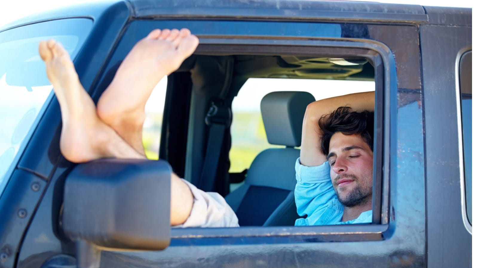A man relaxing in his truck with his feet out the driver's side window to represent how to sleep in your car on a road trip.