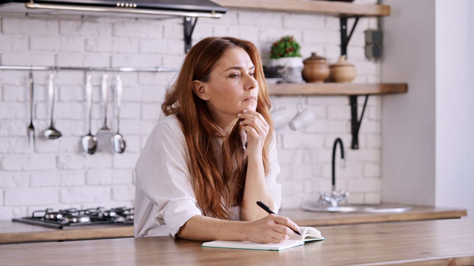 A stressed looking woman in the kitchen with a notebook. She looks like she's trying to remember everything she needs to do, which represents the mental load.