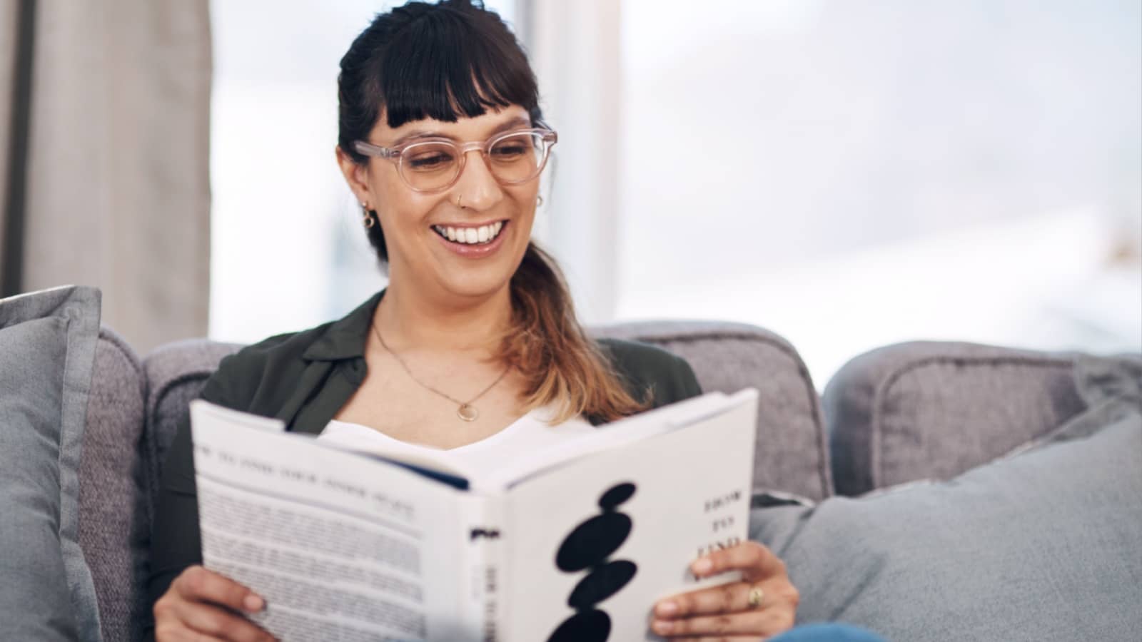 A smiling woman reads a book while sitting on her couch to represent books on happiness.