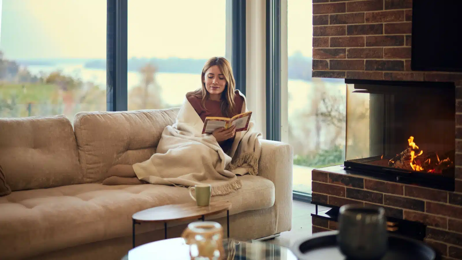 A woman lays on the couch, reading a book next to the fireplace.