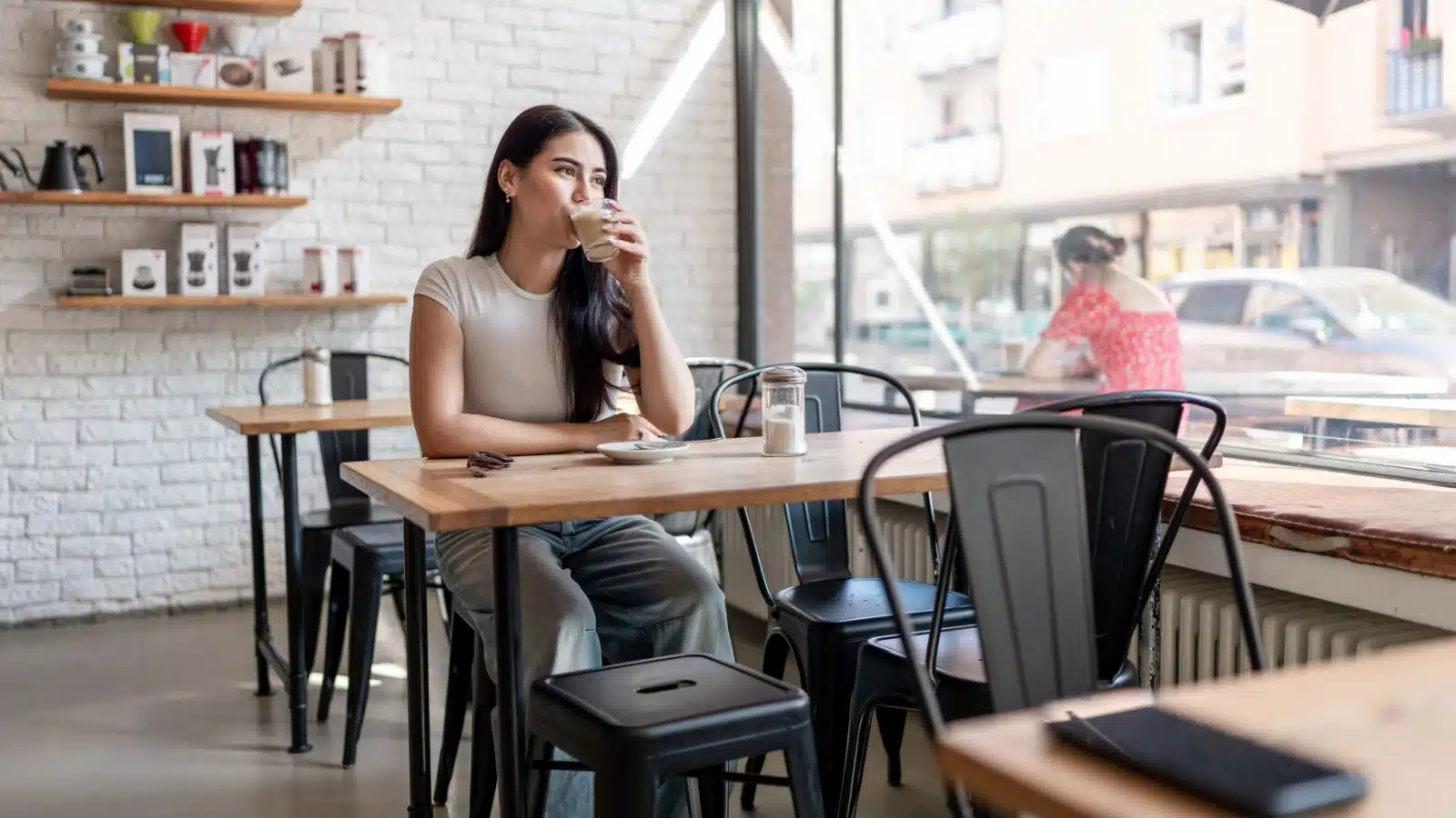 A woman sips coffee while people watching at a cafe with giant windows.
