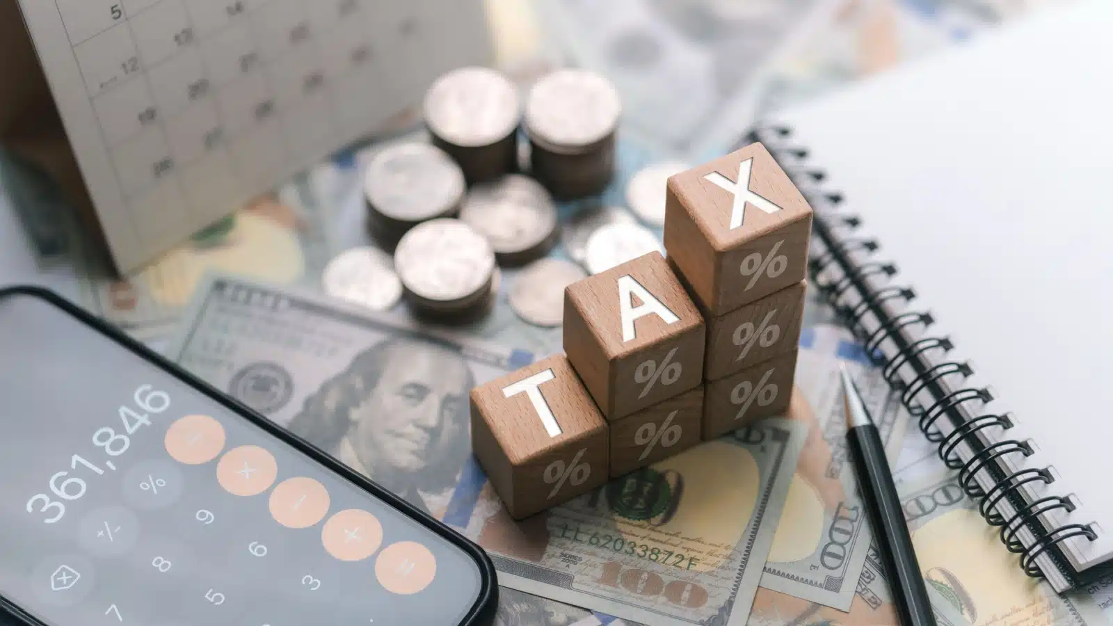 Concept photo representing a tax increase. The word "tax" is spelled out in wooden blocks resting atop money, but the T is lower than the A, which is lower than the X, which represents rising taxes.