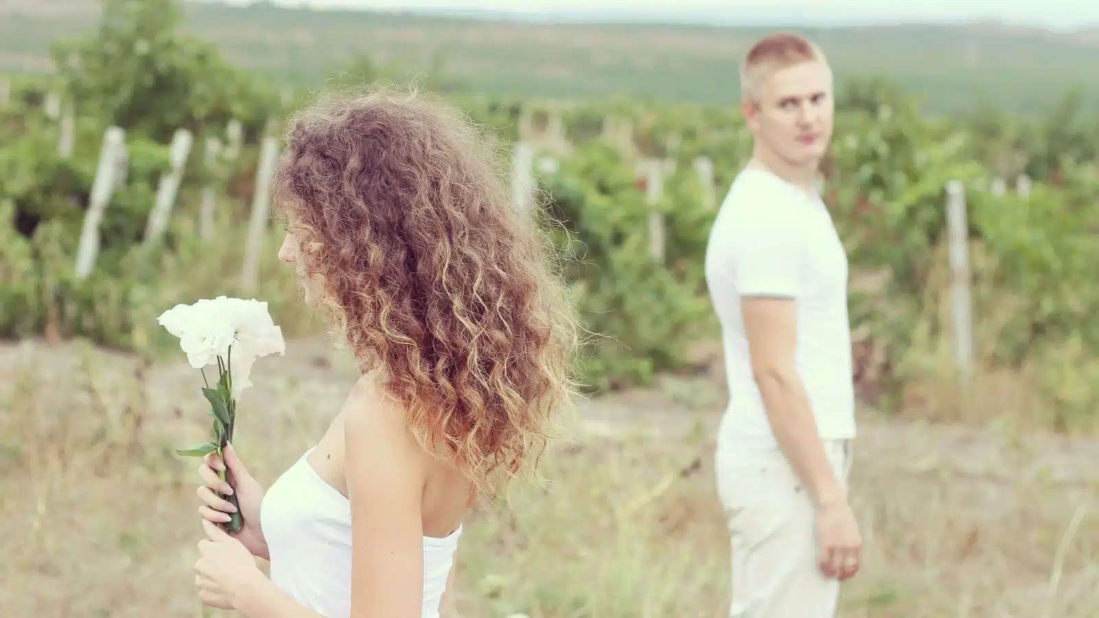 A couple in a field stands apart. The woman smells flowers, ignoring the man, while he looks at her, to represent different dating standards between men and women.