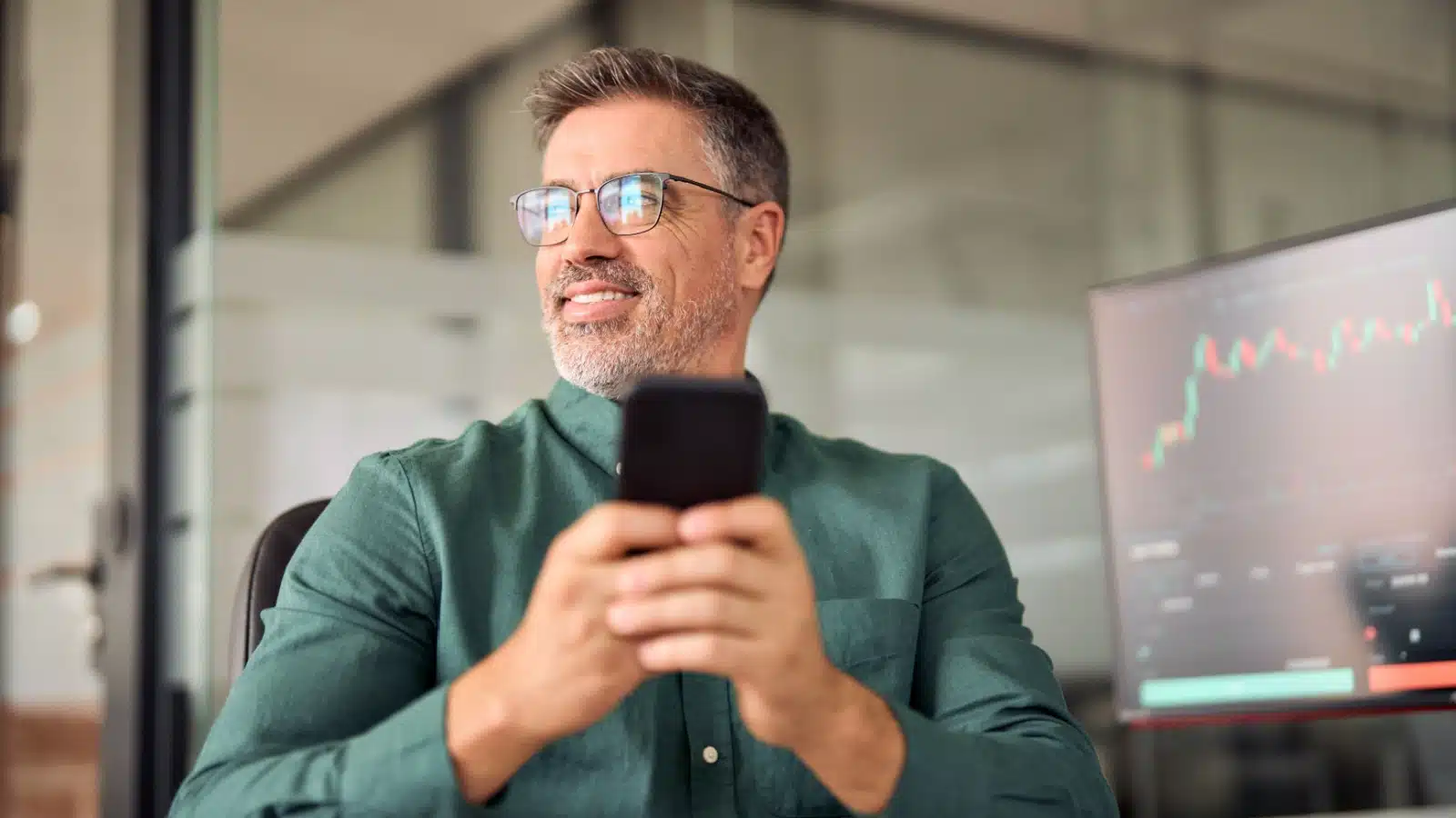 A happy investor uses his phone, while his computer shows investment gains behind him.