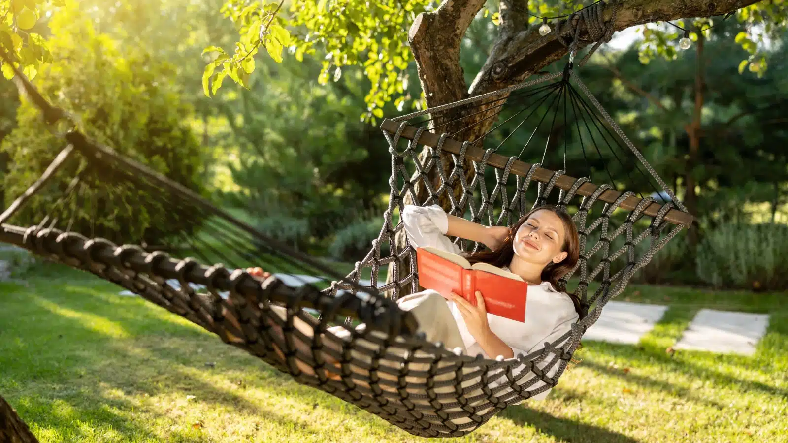 A woman reads a book while resting in a hammock.