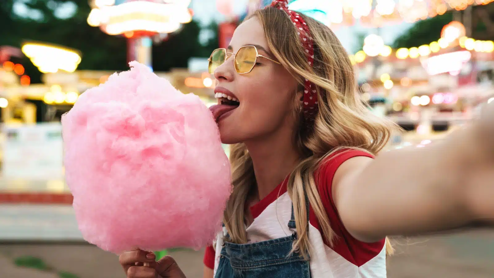 A young woman takes a selfie while enjoying cotton candy at a summer carnival.
